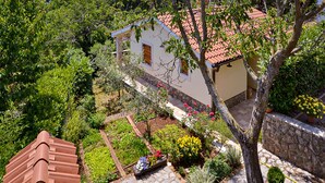 Ein Haus mit rotem Ziegeldach, umgeben von üppiger Vegetation und einem Garten mit verschiedenen Pflanzen und Blumen.