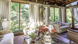 Une pièce chaleureuse avec un plafond en bois, de grandes fenêtres, un canapé, un fauteuil à motifs et une table sur laquelle est posé un vase rempli de fleurs rouges.