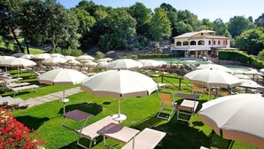 Un área de estar al aire libre bien cuidada, con numerosas sombrillas blancas y sillones de descanso, rodeada de exuberante vegetación y un edificio grande al fondo.