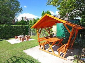 A wooden outdoor seating area with a table and benches, surrounded by a hedge and a fence.
