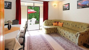 A living room with a sofa, a dining table, and chairs. There is a window with red curtains and a view of greenery outside.