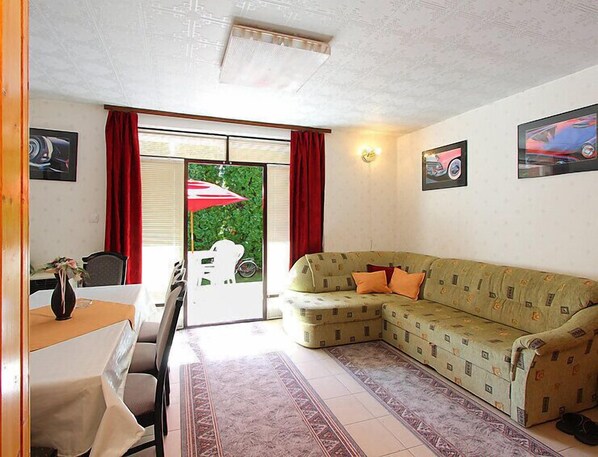 A living room with a sofa, a dining table, and chairs. There is a window with red curtains and a view of greenery outside.