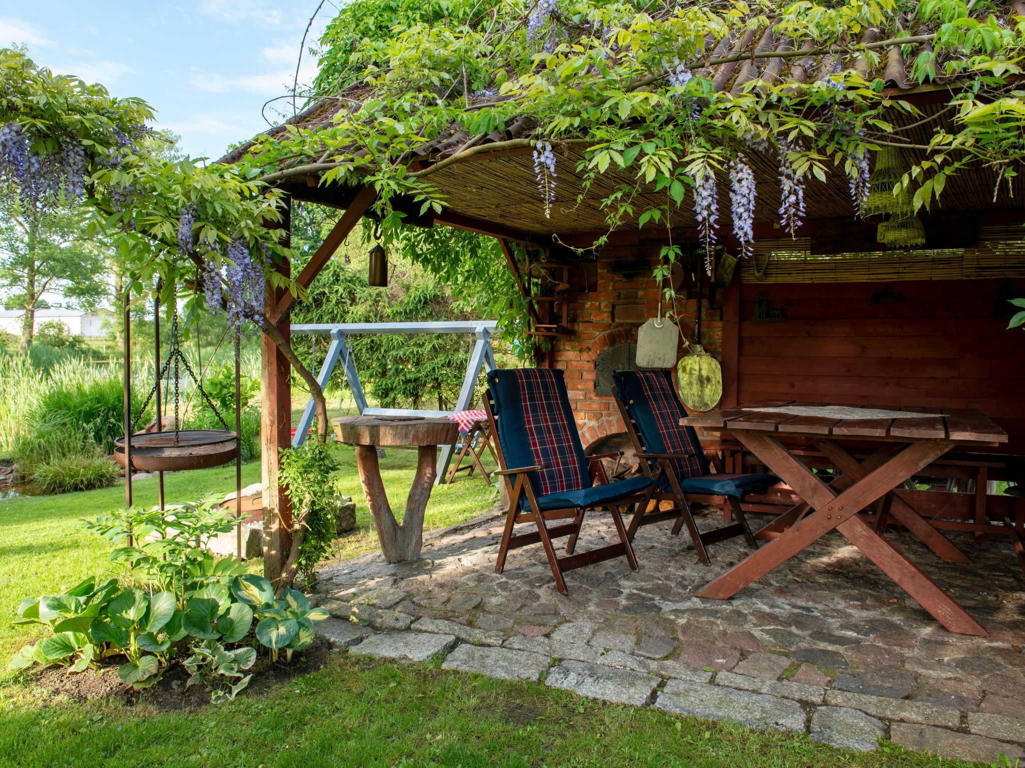 Et hyggeligt udendørs siddeområde med en gyngestol, et træbord og stole under et pergola overdækket med slyngplanter.