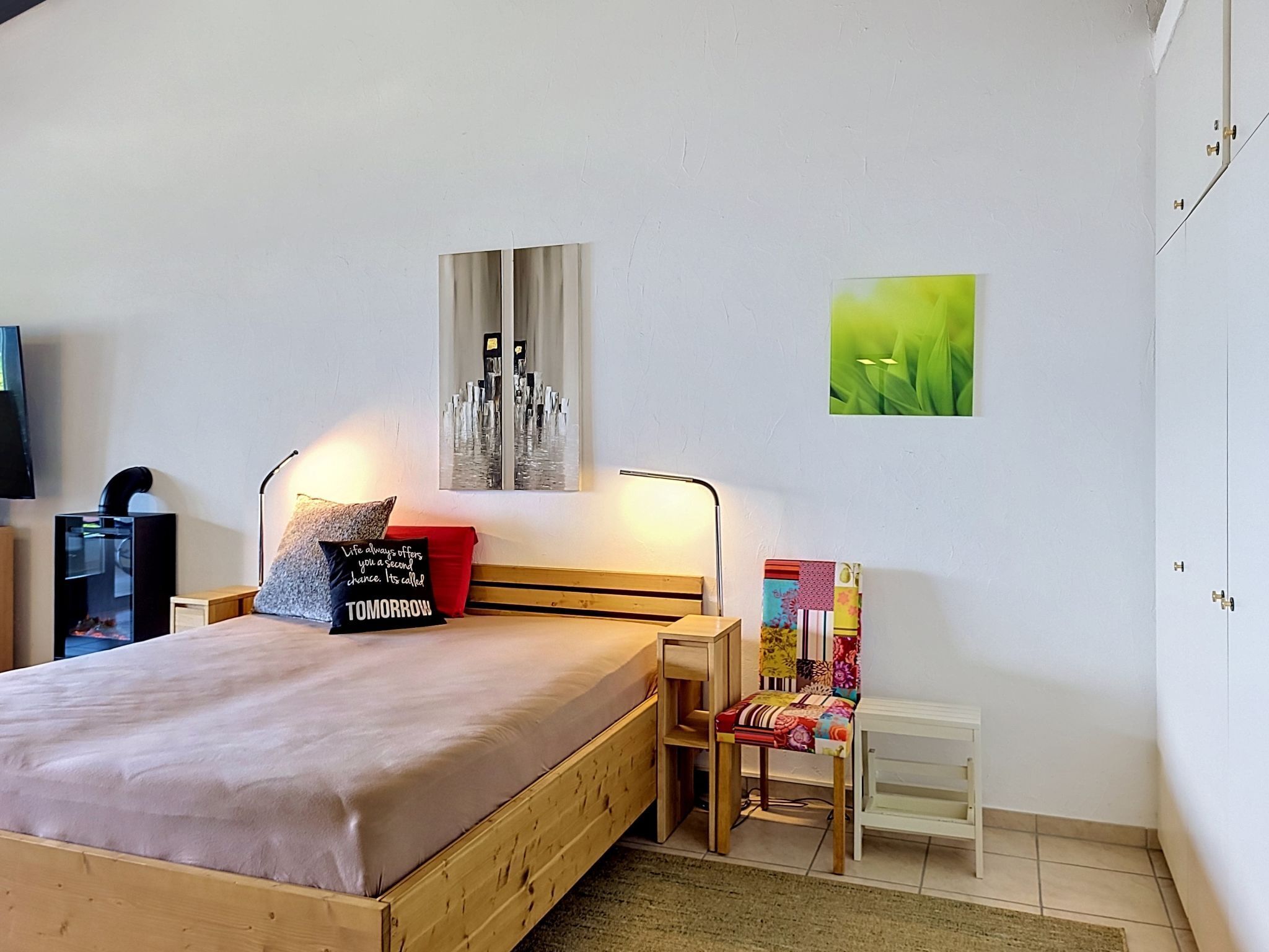 Un dormitorio con cama, mesita de noche, silla y obras de arte en las paredes.