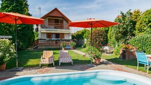 Área de piscina com uma casa ao fundo, dois guarda-sóis vermelhos e uma churrasqueira de tijolos.
