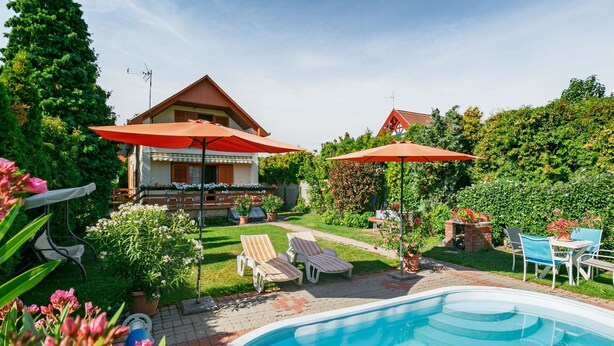 Piscina com espreguiçadeiras e guarda-sóis, rodeada por um jardim e uma casa com varanda.