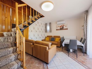 A living room with a wooden staircase, a sofa set, a dining table with chairs, and an air conditioning unit.