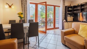 Uma sala de estar moderna com sofá, área de jantar com cadeiras e mesa, televisão e porta de vidro deslizante que dá para um pátio externo.