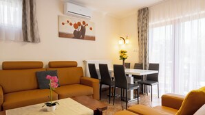 Uma sala de estar com sofá, mesa de centro com planta, área de jantar com cadeiras e um quadro na parede.