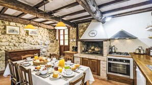 Une cuisine rustique avec un mur en pierre, des poutres en bois, un poêle et un coin repas où une table est dressée pour le petit-déjeuner.