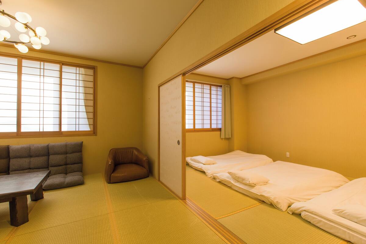 A traditional Japanese room with tatami flooring, a low table, a sofa, and a bed with white bedding.