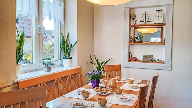 A dining area with a wooden table set for four, potted plants, and a wall-mounted shelf with decorative items.