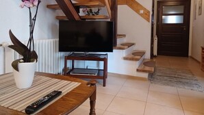 A living room with a wooden staircase, a flat-screen TV on a stand, a dining table with a remote control, and a potted plant.