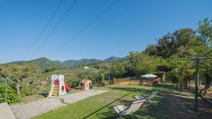 A playground with a slide and swings, surrounded by a grassy area and trees, with mountains in the background. - La Casa al Monte by Interhome (Camaiore)