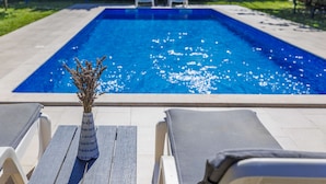 Una piscina rectangular con agua azul cristalina, rodeada por una terraza con baldosas. Hay sillas de descanso y una mesita con un jarrón de flores secas en primer plano.