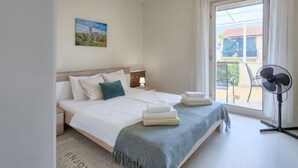 Un dormitorio con cama, mesita de noche, lámpara, ventilador y ventana con cortinas.