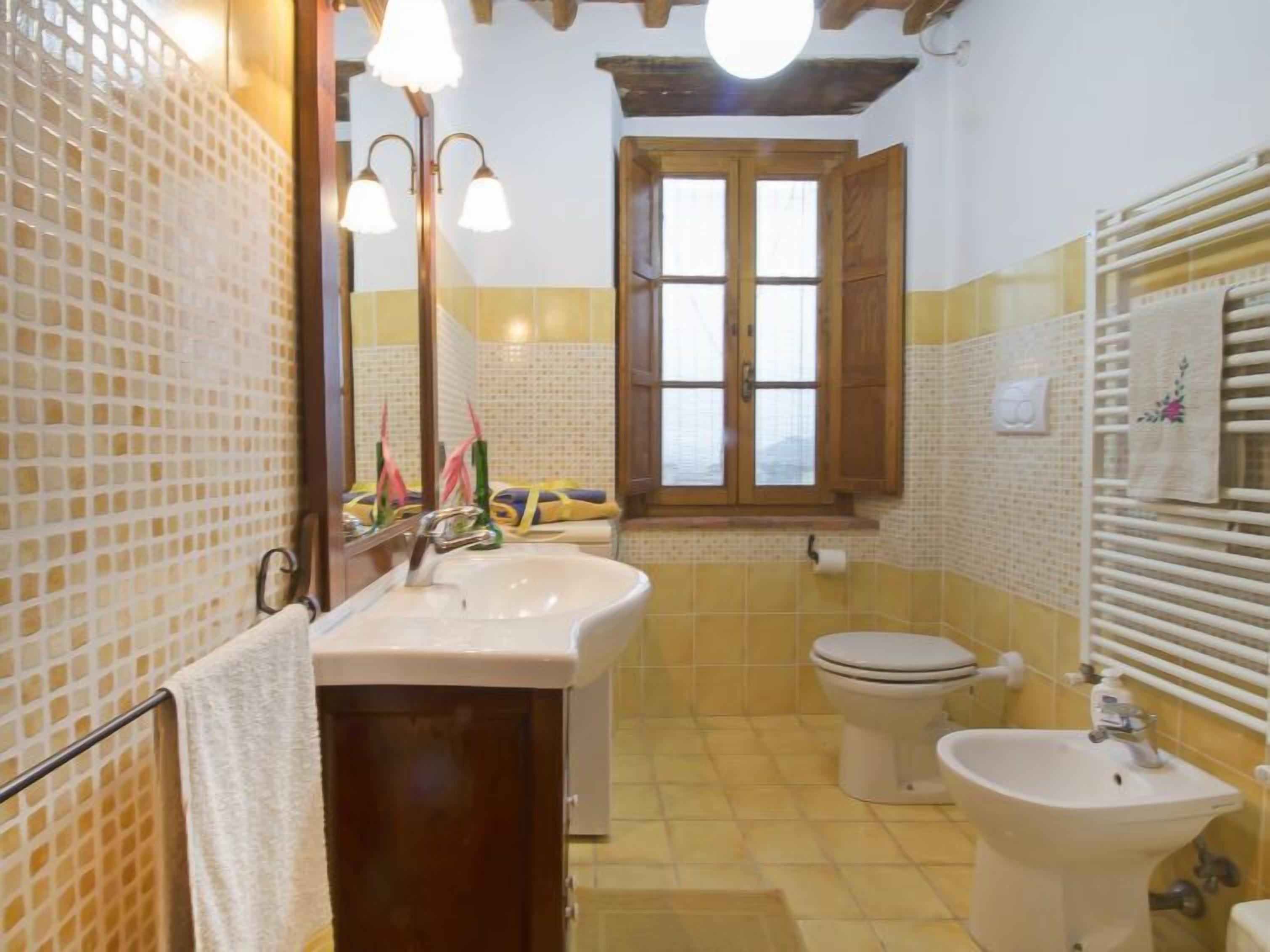 A bathroom with a sink, toilet, and bidet, wooden cabinets, a window, and a towel rack.