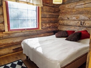 A log cabin bedroom with a bed, pillows, a window with blinds, and a wooden floor.