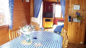 Interior de uma cabana de madeira com uma mesa de jantar, cadeiras, uma televisão e uma estante de livros.