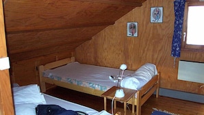 Um pequeno quarto de madeira com uma cama de solteiro, uma escrivaninha com luminária e uma janela com cortinas.