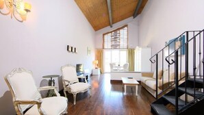 A spacious living room with a wooden floor, a sofa, two armchairs, a small table, and a staircase with a metal railing.