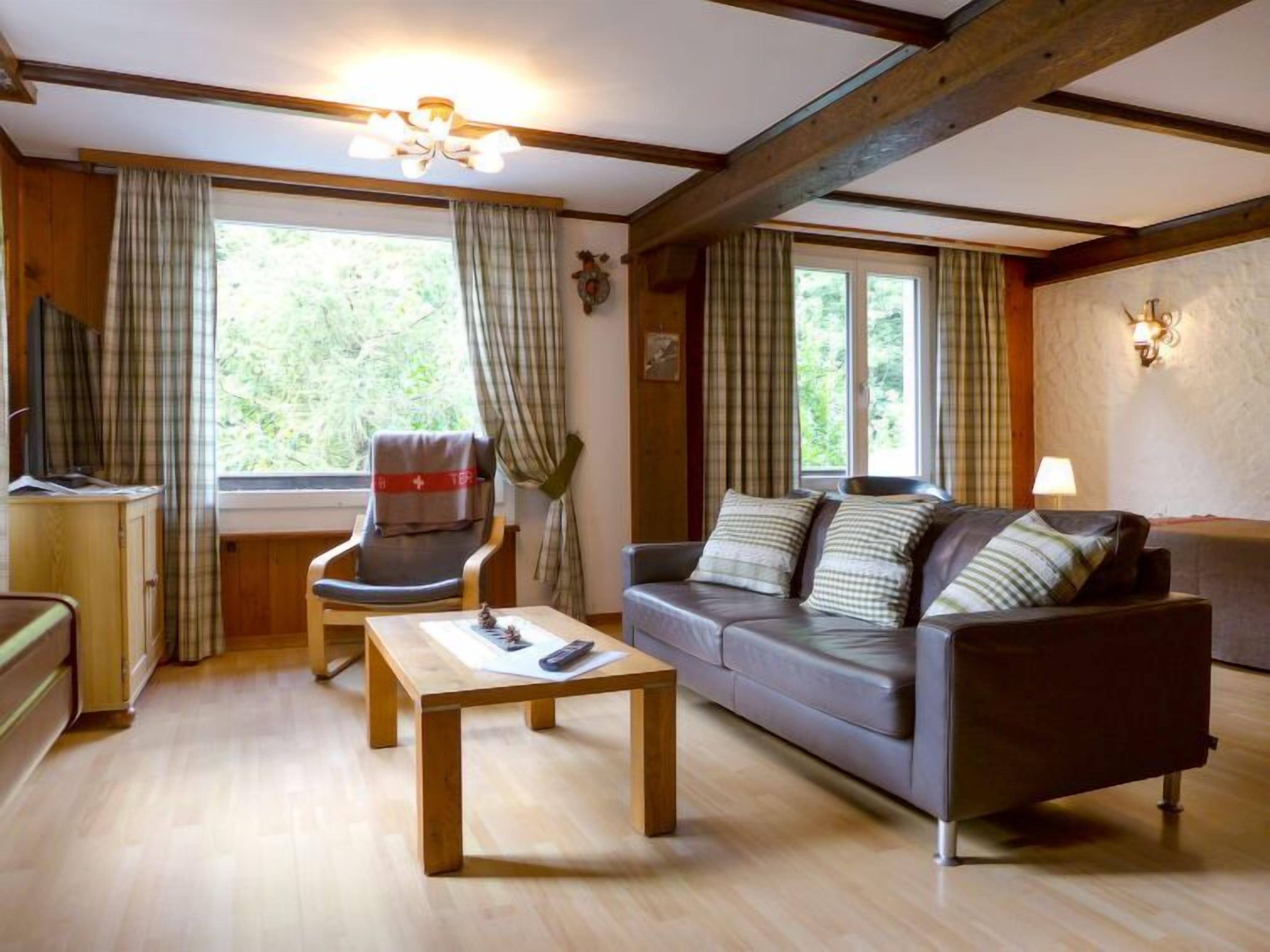 Un salón acogedor con un sofá, un sillón y una mesa de centro de madera. Hay cortinas en las ventanas y una lámpara en la pared.