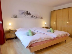 A double bed with a pink and white duvet, two bedside tables with lamps, a wooden wardrobe, and a mountain landscape painting on the wall.