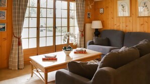 Een gezellige woonkamer met houten wanden, een grijze bank, een houten salontafel met boeken en een raam met gordijnen.