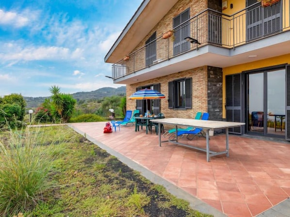 Ein zweistöckiges Haus mit einer Terrassenfläche, auf der sich ein Tisch und Stühle befinden, ein roter Rasenmäher und ein Blick auf die umliegende Landschaft.