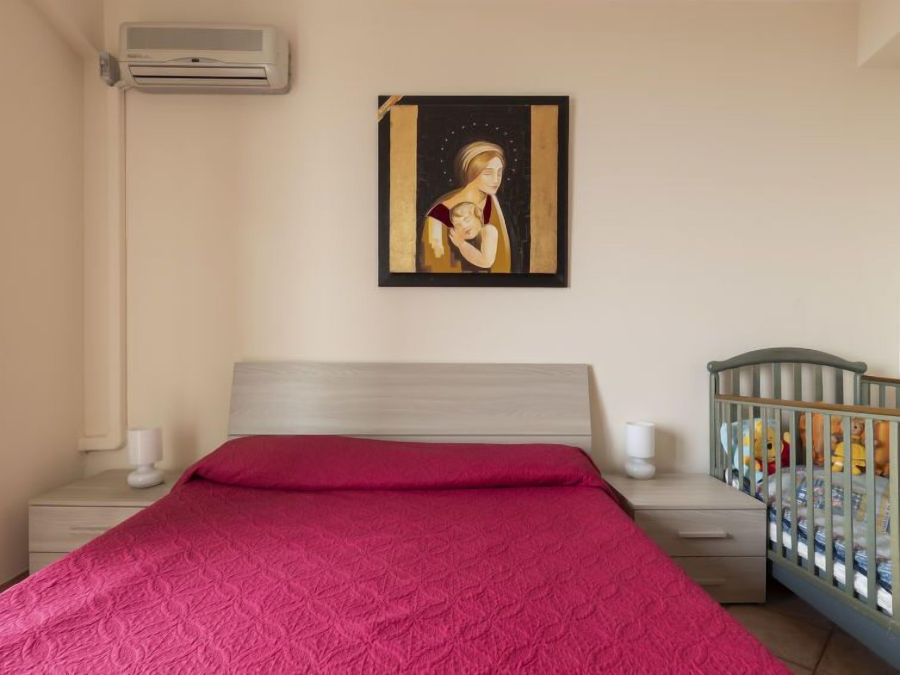 A bedroom with a bed, a crib, a wall-mounted air conditioner, and a framed painting.