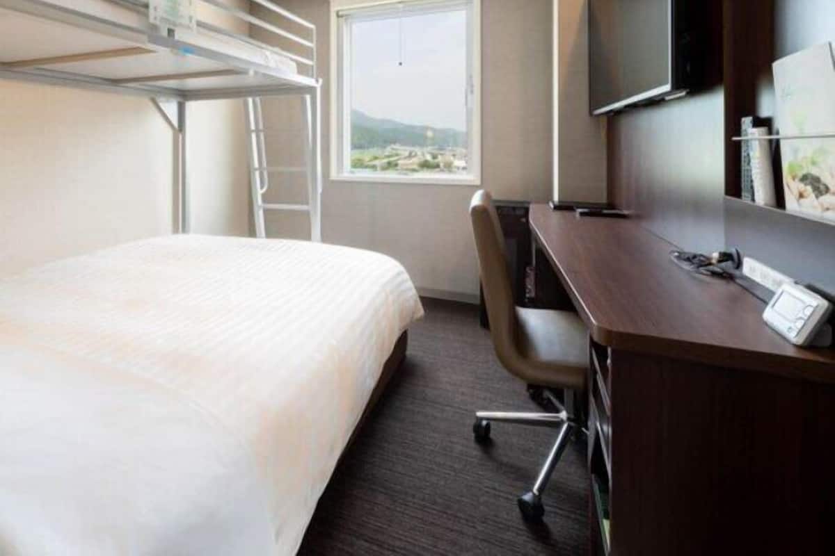 A hotel room with a bunk bed, a desk with a chair, a TV mounted on the wall, and a window with a view.