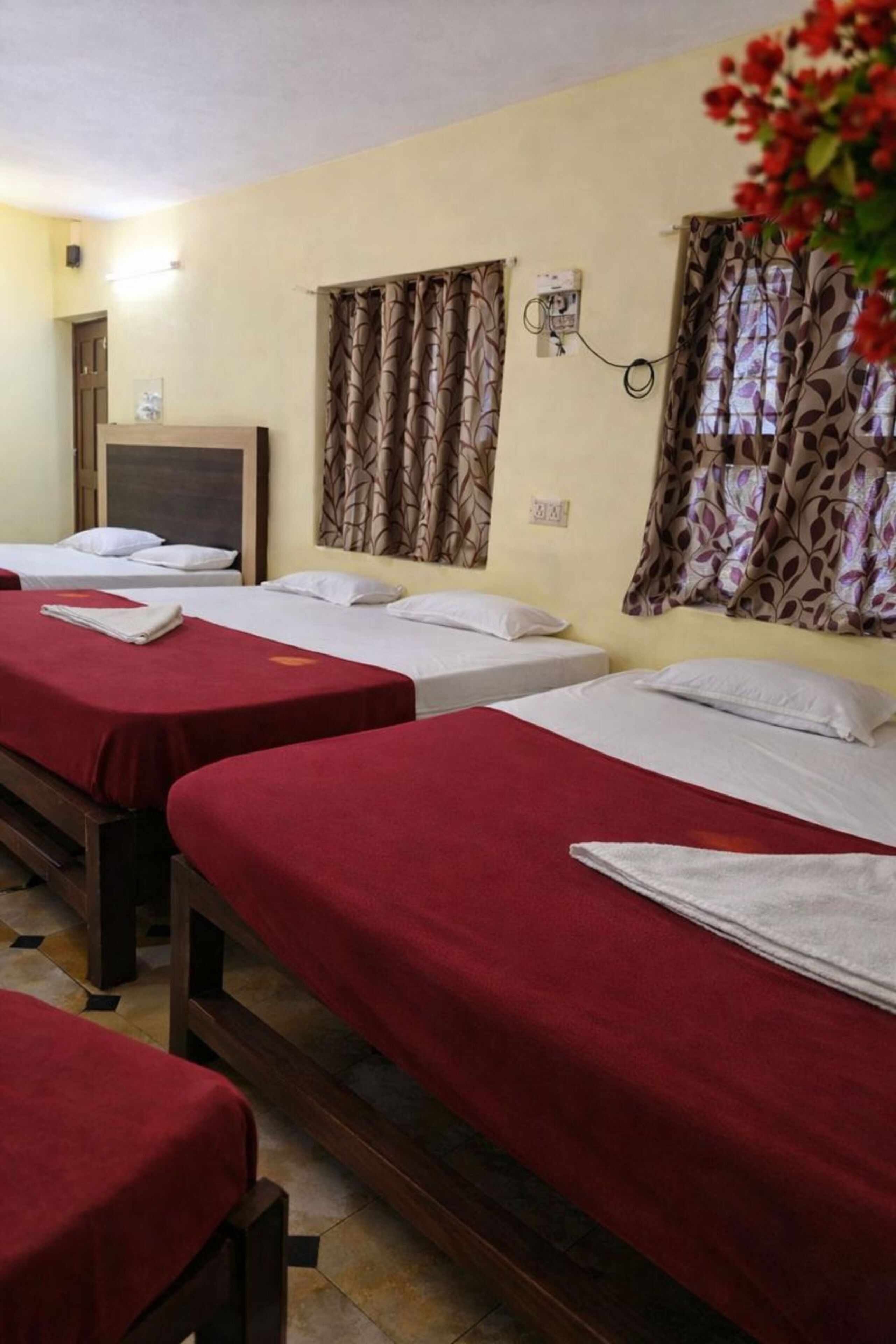 A hotel room with three beds, each with a red bedspread and white pillows. There is a television on a wooden stand, and the room has patterned curtains.