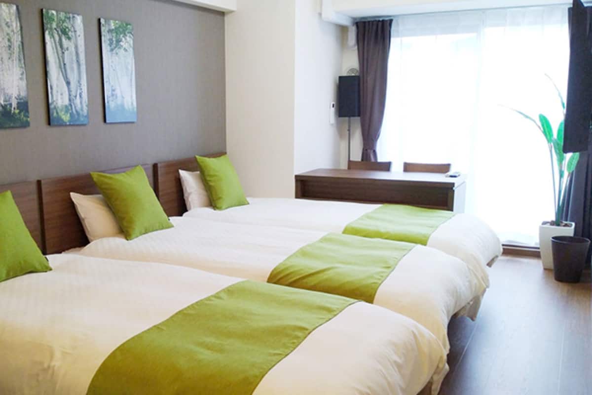 Habitación de hotel con dos camas, ropa de cama verde y blanca, una mesita, un televisor y una ventana con cortinas.