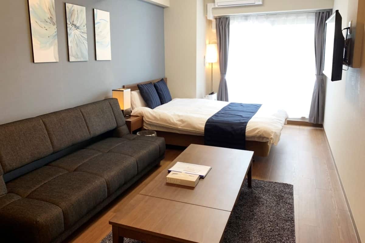 Habitación de hotel con sofá, cama, mesita de noche, lámpara y ventana con cortinas.