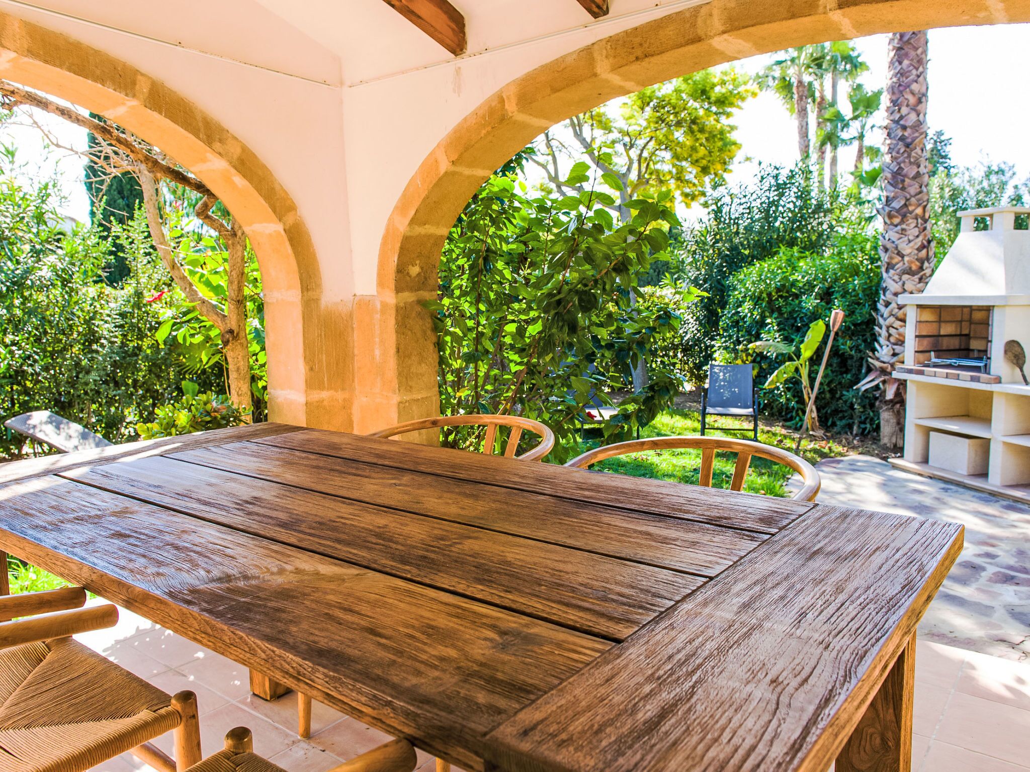 A wooden table with chairs under a stone archway, surrounded by lush greenery and a barbecue area.