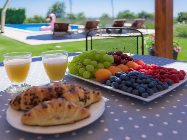 A table with croissants, a fruit platter, and two glasses of juice, set outdoors with a pool and flamingo in the background.