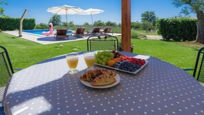 A table with a polka dot tablecloth, a plate of croissants, and a fruit platter with grapes, berries, and oranges. There are two drinks on the table. In the background, there is a pool with lounge chairs and umbrellas. - Vacation home Blog 2 by Interhome (Poreč/Sv. Lovreč)