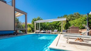 Une piscine dotée de chaises longues et d’une pergola, le tout entouré de verdure.
