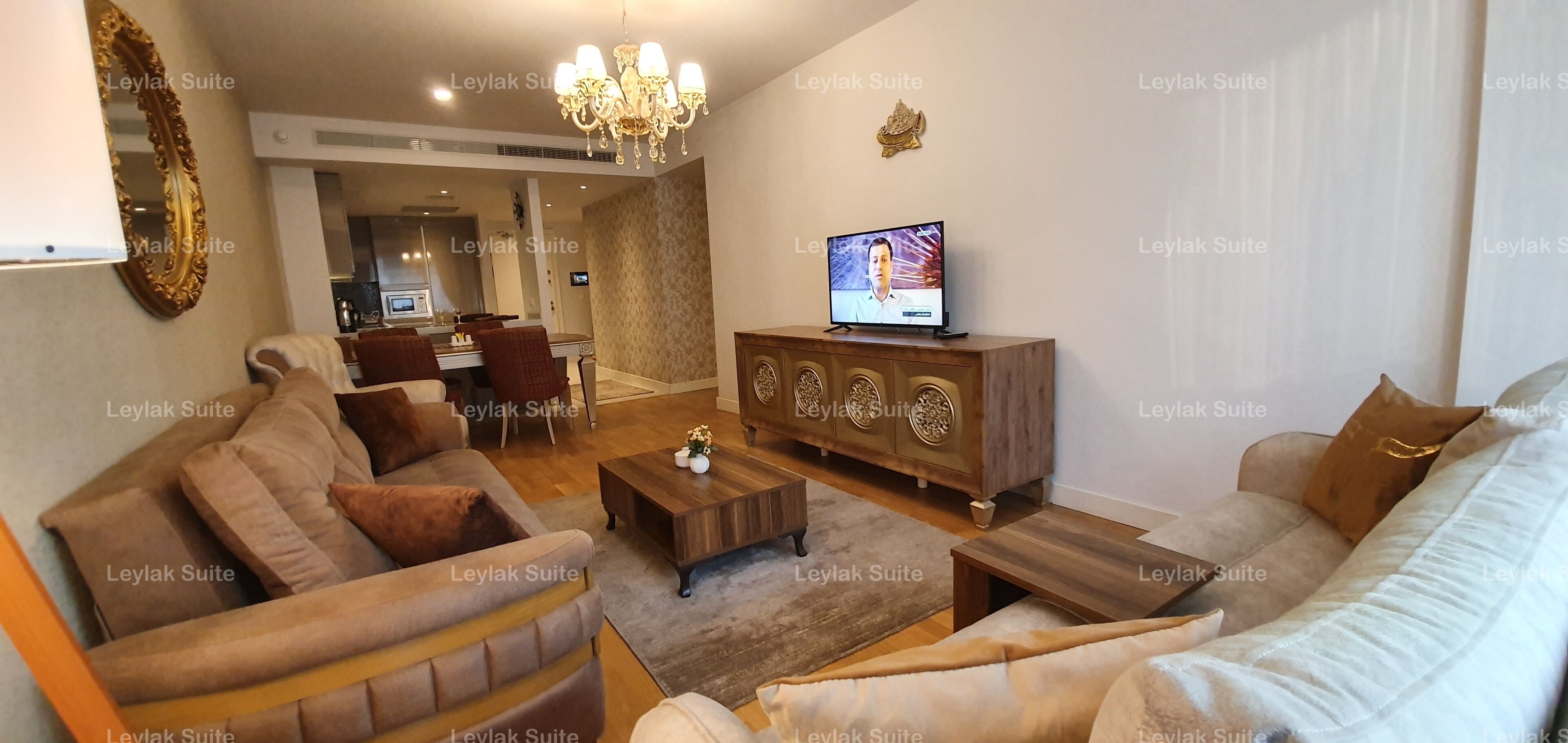 Un salon comprenant un canapé, des fauteuils, une table basse, un téléviseur posé sur un meuble en bois et un lustre.