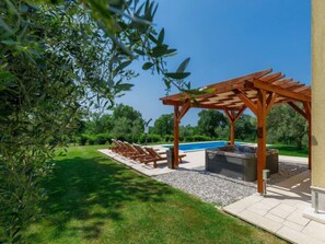 Une pergola en bois avec des fauteuils de jardin et un barbecue, une piscine et une pelouse bien entretenue.