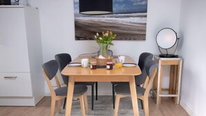 A dining area with a wooden table, four chairs, a vase with flowers, and a painting of a beach scene. - Gezeiten by Interhome (Norddeich)