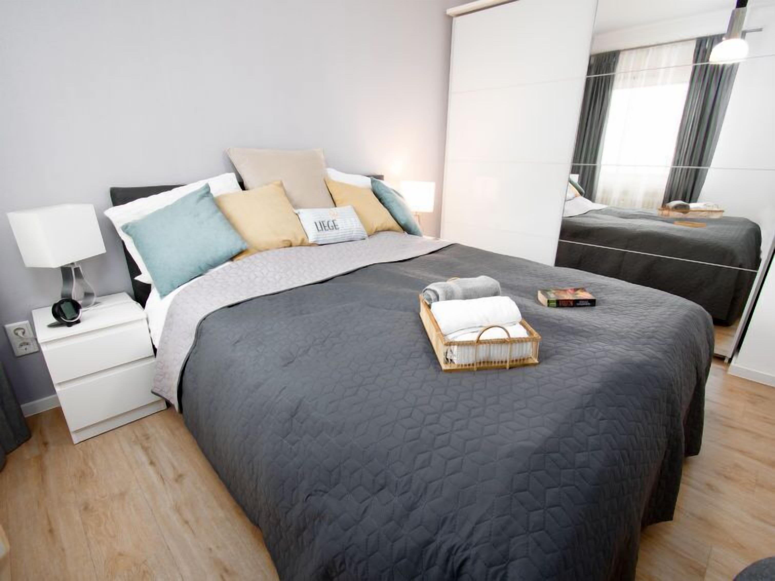A modern bedroom with a large bed, a smaller bed, a bedside table with a lamp, and a bedside basket.