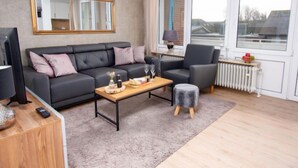 A modern living room with a black sofa, a wooden coffee table, a grey ottoman, and a large window with a view of buildings. - Gezeiten by Interhome (Norddeich)