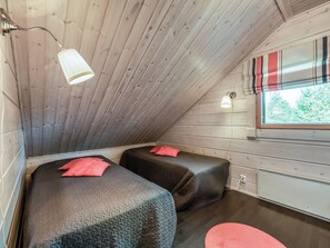A small, cozy room with wooden walls and a sloped ceiling. There are two beds with dark bedding and red pillows. A window with striped curtains allows natural light in.