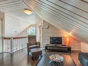 A cozy attic room with a wooden sloped ceiling, a comfortable armchair, a flat-screen TV, and a decorative table.