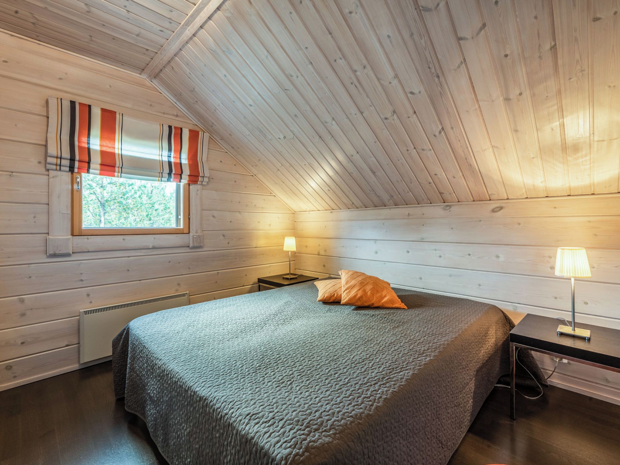 Une chambre avec un plafond en pente, un lit recouvert d’une couverture à motifs, deux lampes de chevet et une fenêtre avec des rideaux à rayures.