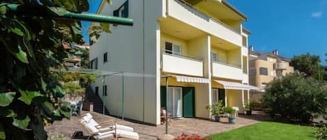 A two-story building with a balcony, a small garden with a lawn and flowers, and a lounge chair.