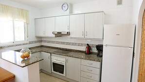 A compact kitchen with white cabinets, a built-in oven, a refrigerator, and a clock on the wall. - Vacation home Vicky by Interhome (Deltebre)