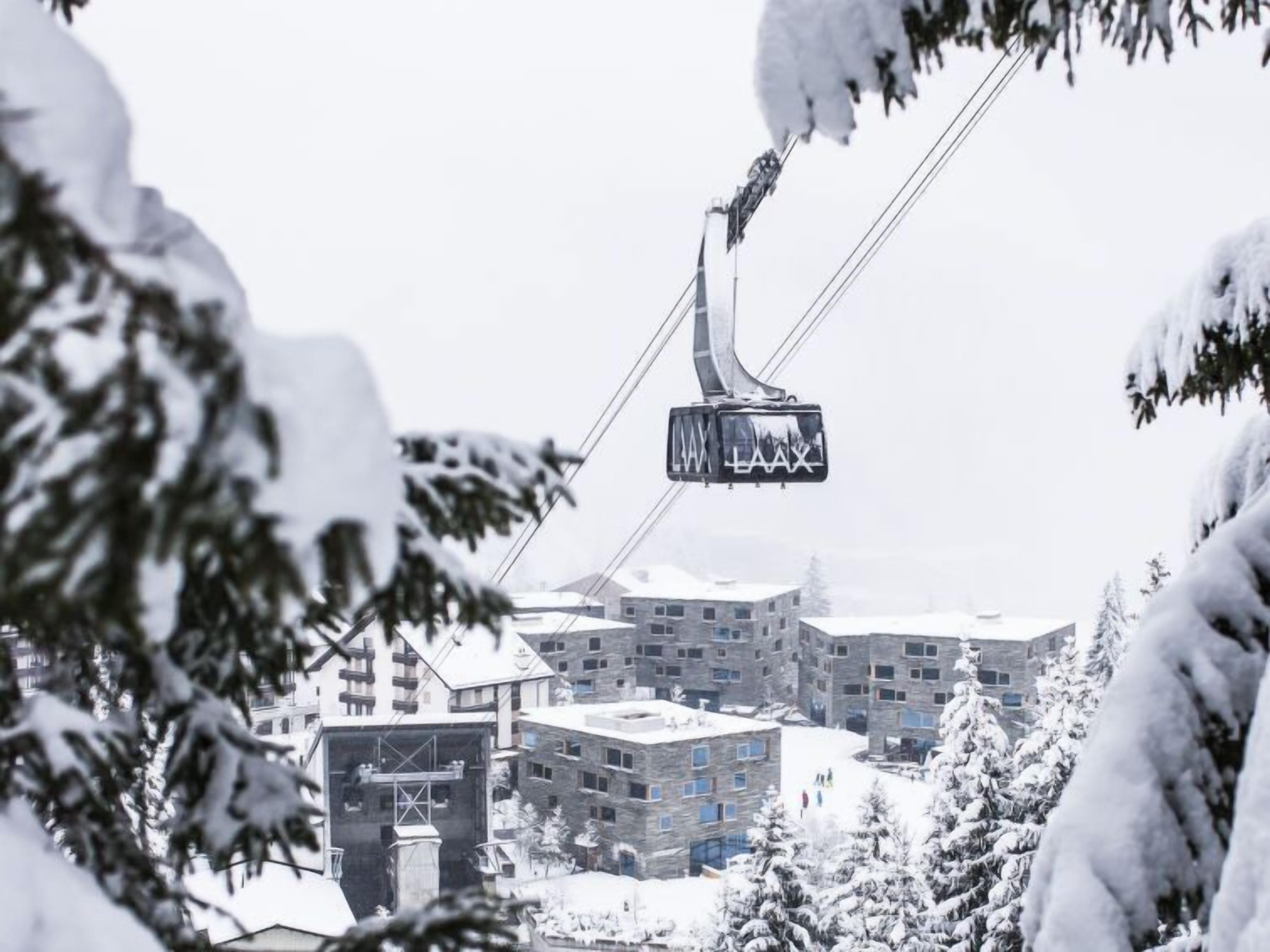Un télésiège avec des cabines suspendues à des câbles, entouré de bâtiments et d’arbres recouverts de neige.