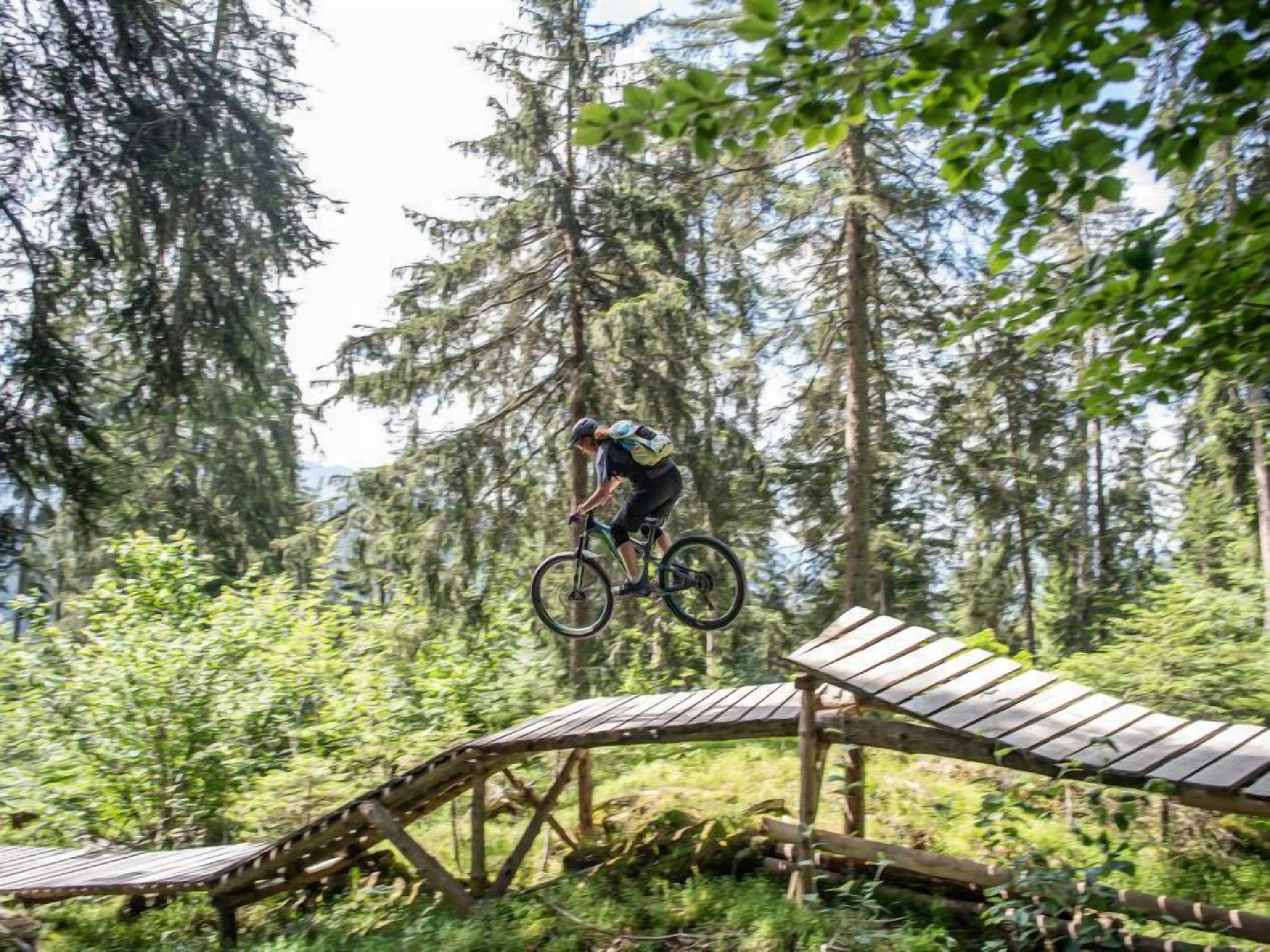 Um ciclista de montanha realizando um salto sobre uma ponte de madeira numa área arborizada.